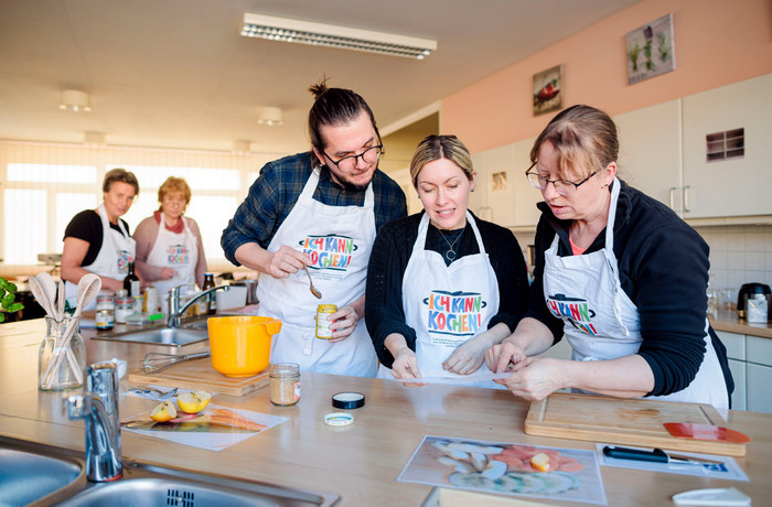 Foto: Sarah Wiener Stiftung I photothek Drei Fortbildungsteilnehmende stehen in der Übungsküche an der Arbeitsplatte. Vor ihnen liegt ein Rezept, dass sie gemeinsam studieren.