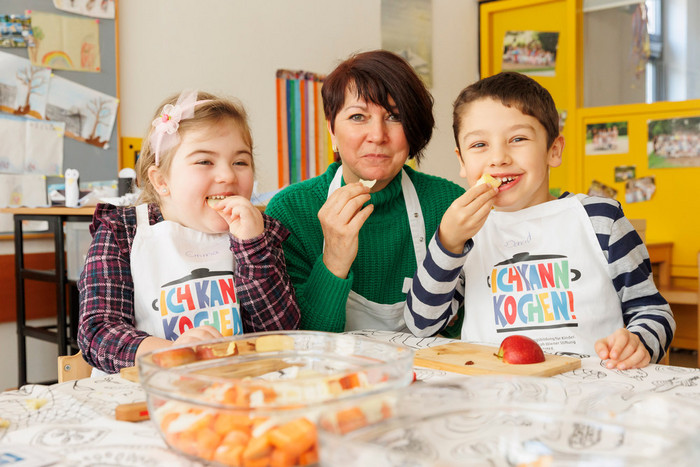Foto: Sarah Wiener Stiftung I photothek Eine Kitamitarbeiterin sitzt mit zwei Kindern am Tisch in der Kita. Sie essen Äpfel und schauen lächelnd in die Kamera.