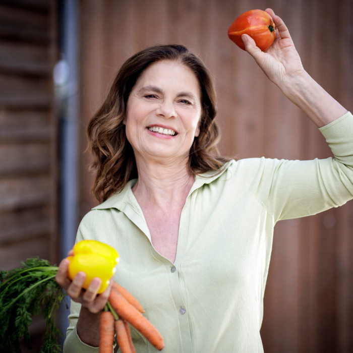 Foto: Sarah Wiener Stiftung I photothek Die Stiftungsgründerin Sarah Wiener posiert mit Obst und Gemüse in beiden Händen lächelnd für die Kamera.